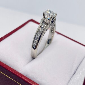 May include: A white gold ring with a large, clear center stone and smaller stones set along the band. The ring is displayed inside a red jewelry box with a white interior. The ring is marked with "14K".
