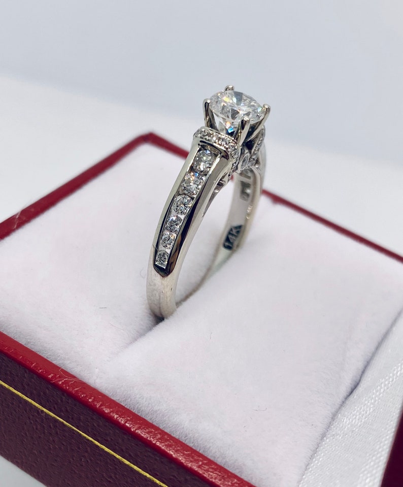 May include: A white gold ring with a large, clear center stone and smaller stones set along the band. The ring is displayed inside a red jewelry box with a white interior. The ring is marked with "14K".