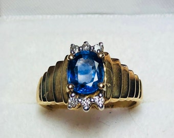 Könnte beinhalten: Ein goldener Ring mit einem großen, oval geschliffenen blauen Edelstein als Mittelpunkt. Der Ring ist mit kleinen, klaren Edelsteinen verziert und hat ein geripptes Design am Band. Der Ring ist vor einem weißen Hintergrund platziert.