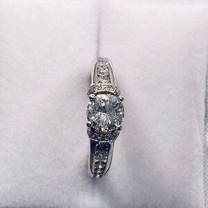 May include: A silver engagement ring with a large, round, clear center stone. The band is adorned with smaller, clear stones. The ring is presented in a red-lined white box, creating a striking contrast.