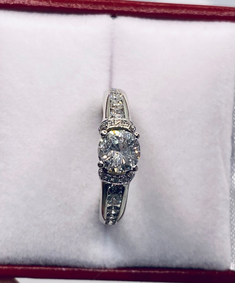 May include: A silver engagement ring with a large, round, clear center stone. The band is adorned with smaller, clear stones. The ring is presented in a red-lined white box, creating a striking contrast.