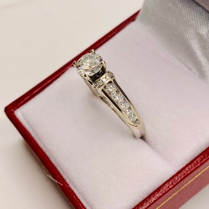 May include: A diamond engagement ring with a large, round-cut center stone. The ring features a white gold band with smaller diamonds set along the sides. The ring is displayed in a red jewelry box with a white interior.