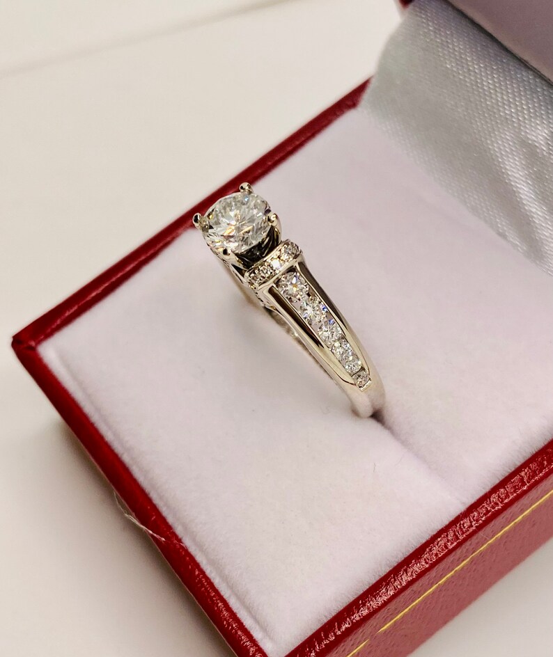 May include: A diamond engagement ring with a large, round-cut center stone. The ring features a white gold band with smaller diamonds set along the sides. The ring is displayed in a red jewelry box with a white interior.