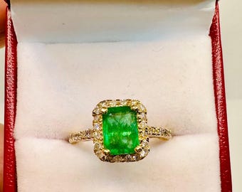 May include: A gold ring with a large, rectangular emerald center stone, surrounded by a halo of smaller diamonds. The ring is set in a red jewelry box with a white interior.