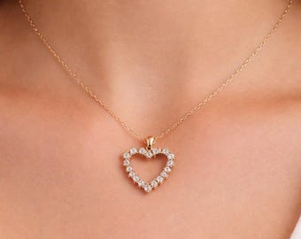 Puede incluir: Un collar dorado con un colgante en forma de corazón. El colgante está adornado con pequeñas piedras preciosas brillantes. El collar se lleva alrededor del cuello, con el colgante descansando sobre el pecho.