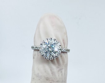 Könnte beinhalten: Ein silberner Verlobungsring mit einem großen runden Diamanten in der Mitte, der von einem Heiligenschein aus kleineren Diamanten umgeben ist. Die Ringschiene ist ebenfalls mit kleineren Diamanten besetzt.