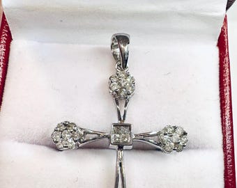 May include: A silver pendant in the shape of a cross, set with multiple small diamonds. The cross is displayed inside a red and white jewelry box, with the pendant's bail at the top.
