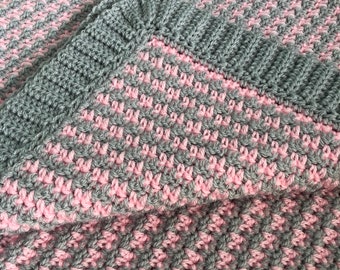 May include: A handmade crochet blanket in grey and pink. The blanket has a textured stitch pattern with a grey border. The soft colours and cosy design make it a perfect addition to any home decor.
