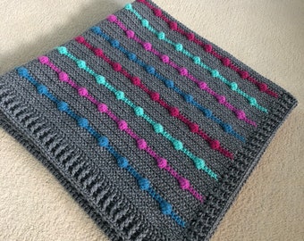 May include: A grey crochet blanket featuring rows of bobble stitches in blue, turquoise, pink, and red. The blanket has a decorative border and is neatly folded. This handmade blanket is ideal for a baby or as a decorative throw.