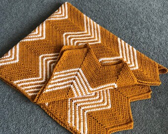 May include: Mustard yellow and white crochet blanket with a geometric chevron pattern. The blanket is folded, showing the detailed stitch work and design. The blanket is made of soft yarn.