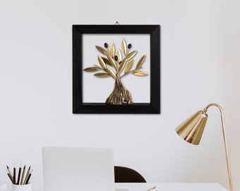 Op de afbeelding: Een ingelijst kunstwerk met een goudkleurige olijfboom met zwarte olijven. De boom staat tegen een witte achtergrond in een zwarte vierkante lijst. Het kunstwerk hangt aan een witte muur boven een bureau.