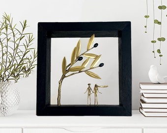 Op de afbeelding: Een zwart ingelijst wandkunstwerk met een gouden metalen olijftak met zwarte olijven en twee gouden metalen figuren die eronder staan.