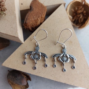 May include: A pair of silver-coloured earrings. Each earring features a stylized floral design with dangling circular charms. The earrings have a hook closure. The background includes a wooden surface and natural elements.