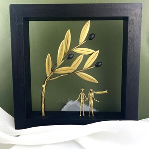 May include: A gold metal olive branch sculpture with black olives and two gold metal figures standing under the branch in a black frame.