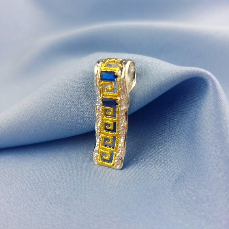 Peut inclure: Un pendentif en argent avec un motif de cl&eacute; grecque dor&eacute;e et une pierre pr&eacute;cieuse bleue. Le pendentif est sur un fond de tissu bleu.