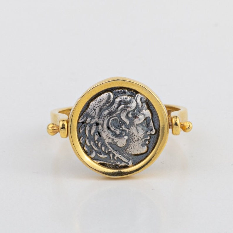 May include: A gold ring with a silver coin featuring a profile of a man's face. The coin is set in a round bezel and the ring has a hinged band.