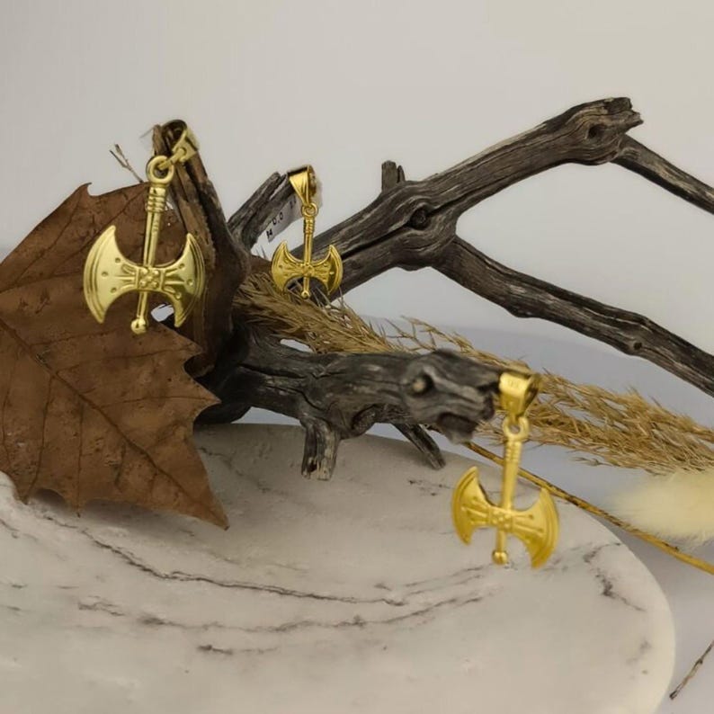 K&ouml;nnte beinhalten: Drei goldfarbene Axt-Anh&auml;nger auf einem Holzast und einer Marmorfl&auml;che. Die Axt-Anh&auml;nger haben ein doppelseitiges Design mit einer Schlaufe oben zum Anbringen an einer Kette.