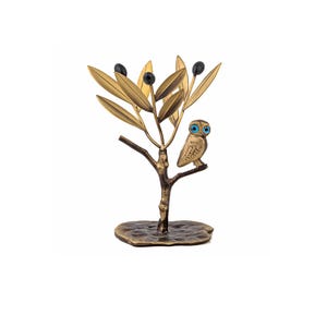 May include: A bronze-toned metal sculpture of an owl perched on a branch of an olive tree. The tree features textured golden leaves and three dark spherical olives. The owl has prominent bright blue eyes and a detailed feathered body.