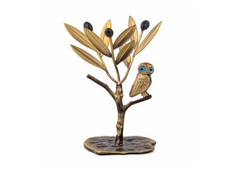 May include: A bronze-toned metal sculpture of an owl perched on a branch of an olive tree. The tree features textured golden leaves and three dark spherical olives. The owl has prominent bright blue eyes and a detailed feathered body.