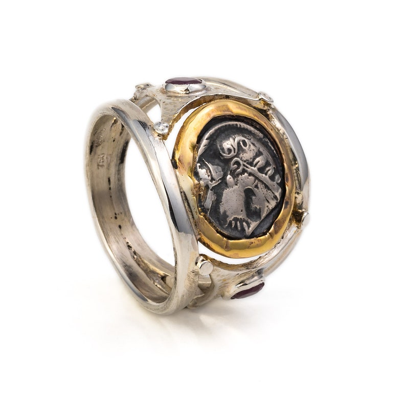 May include: A silver ring with a gold coin set in the center. The coin features a detailed image of a head with a crown. The ring is accented with red gemstones.