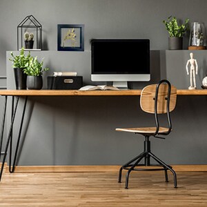 May include: A home office workspace with a black computer monitor, a wooden desk with black hairpin legs, a black office chair with a wooden seat, and a framed print of a gold leaf branch on a gray wall.