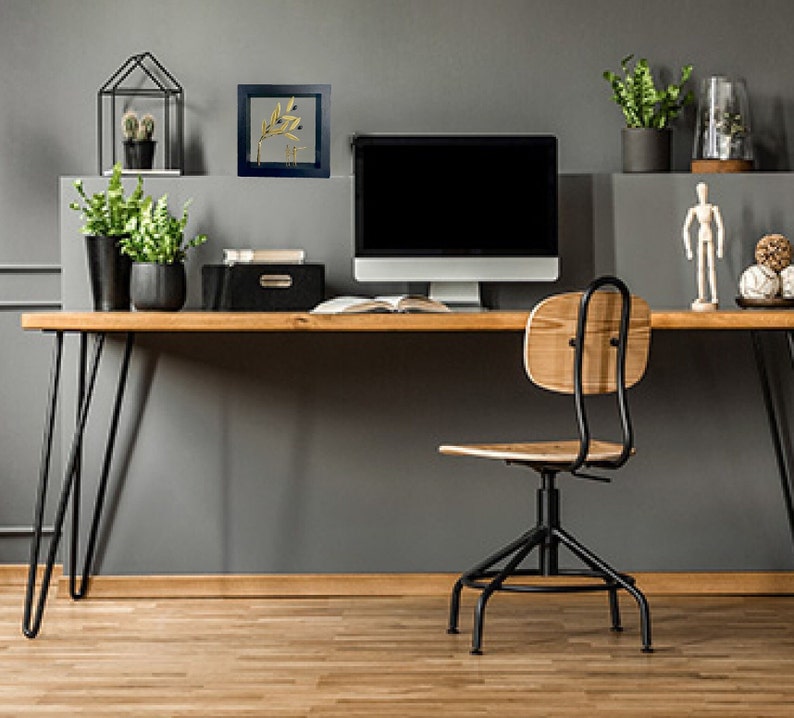 May include: A home office workspace with a black computer monitor, a wooden desk with black hairpin legs, a black office chair with a wooden seat, and a framed print of a gold leaf branch on a gray wall.