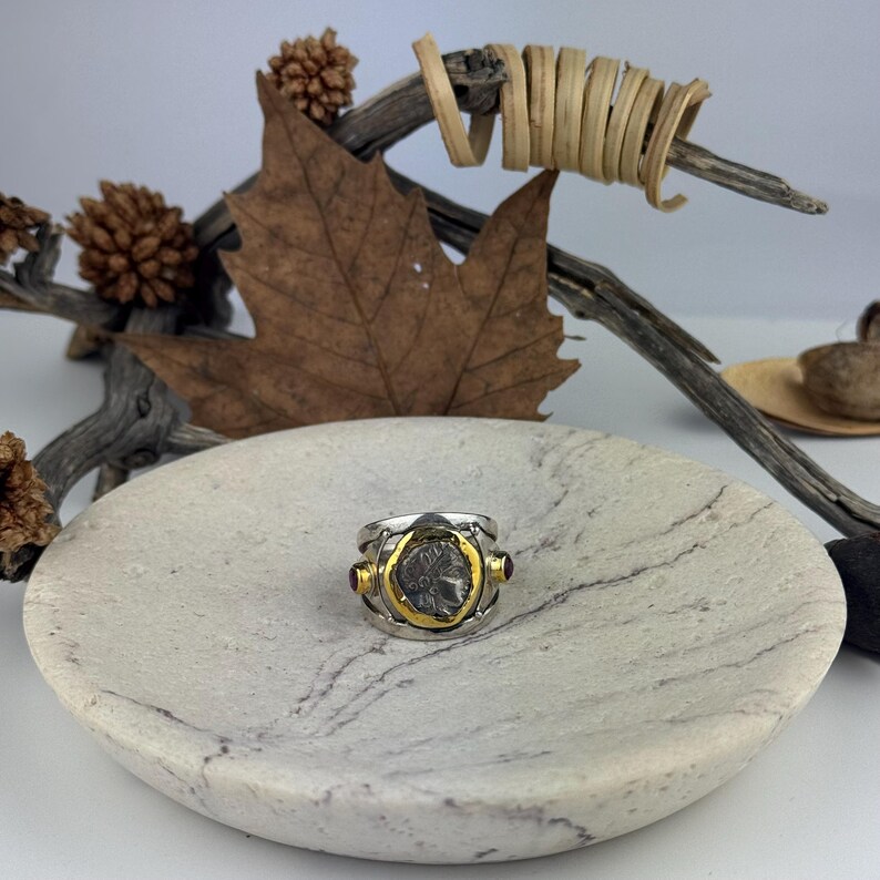 May include: A silver ring with a gold band and a coin-shaped design featuring a profile of a person. The ring is set on a white marble surface.