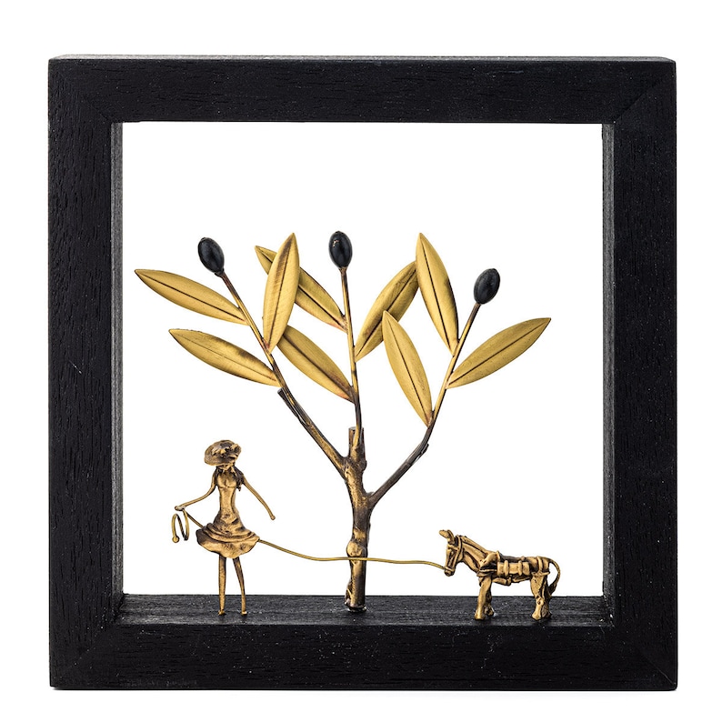 May include: A black framed wall art piece featuring a gold metal olive tree with black olives, a small gold metal figure of a person holding a rope attached to a gold metal donkey.