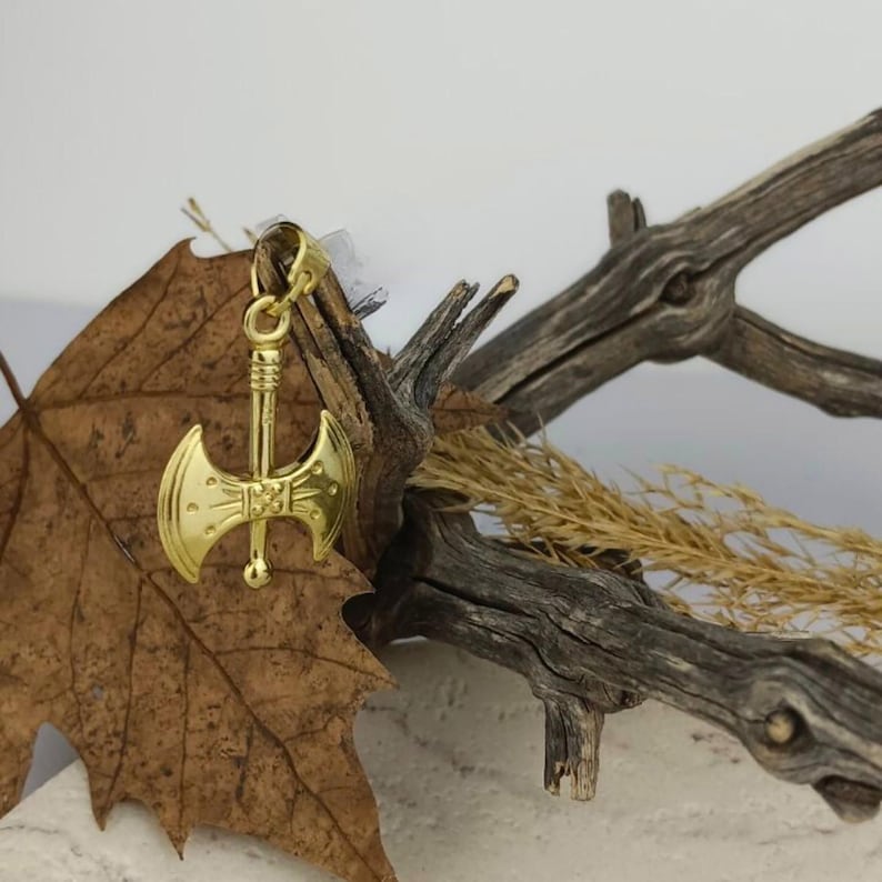 K&ouml;nnte beinhalten: Goldfarbener Doppelkopf-Axt-Anh&auml;nger mit detailliertem Design. Der Anh&auml;nger h&auml;ngt an einem Ast mit einem trockenen Blatt.