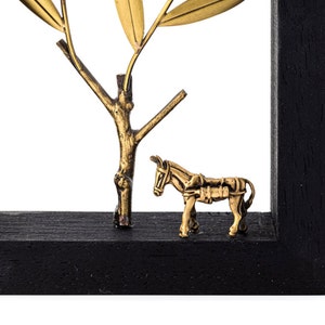 May include: A gold metal sculpture of a donkey standing next to a gold metal olive tree. The sculpture is framed in a black wooden frame.