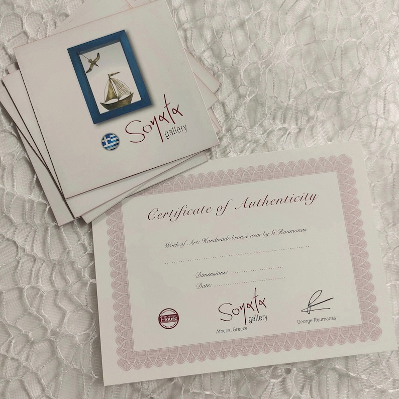May include: Certificate of Authenticity for a handmade bronze item by G. Roumanas. The certificate is printed on white paper with a pink border and features the text "Soyata gallery" and "Athens, Greece".