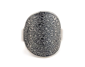May include: A silver ring with a circular design featuring an intricate pattern of symbols and letters. The ring is oval shaped and has a smooth band.