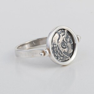 May include: A silver ring with a spinning coin design featuring a detailed profile of a mythical creature. The coin is set in a round bezel and the ring band is plain.