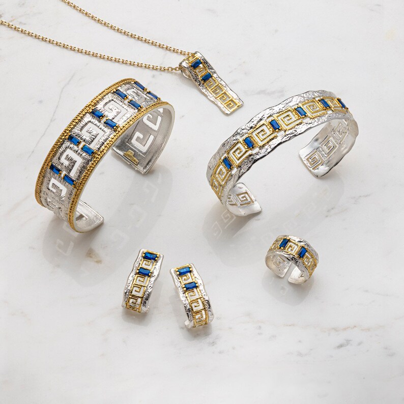 Peut inclure: Un ensemble de bijoux en argent et en or avec un motif de cl&eacute; grecque et des pierres pr&eacute;cieuses bleues. L'ensemble comprend un collier, deux bracelets, une bague et une paire de boucles d'oreilles.