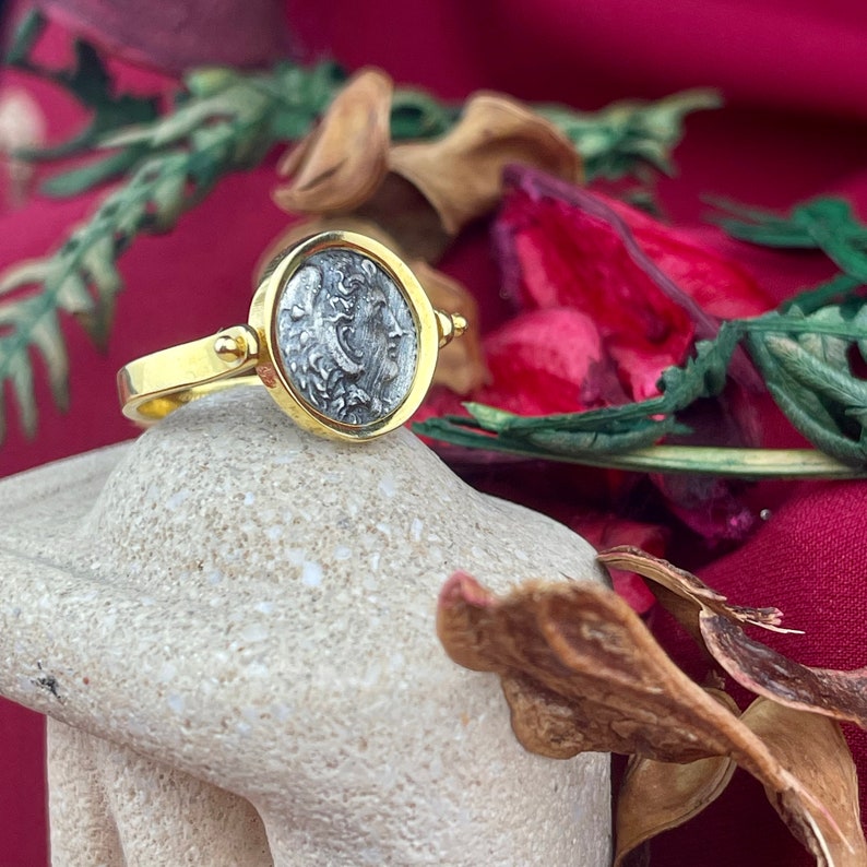 May include: A gold ring with a silver coin featuring a profile of a woman's face. The coin is set in a bezel setting and the ring has a simple band.