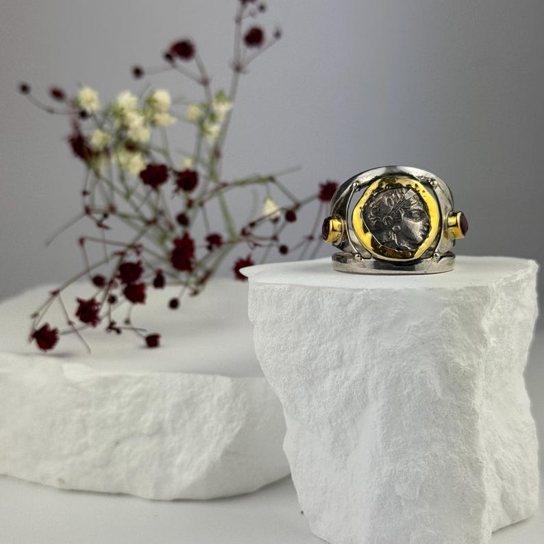 May include: A silver ring with a gold-plated coin featuring a profile of a person's face. The ring is accented with two small, dark red gemstones.