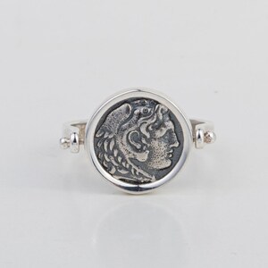 May include: A silver ring with a round coin-shaped design featuring a profile of a person's head with a laurel wreath. The ring is open and has a hinged design.