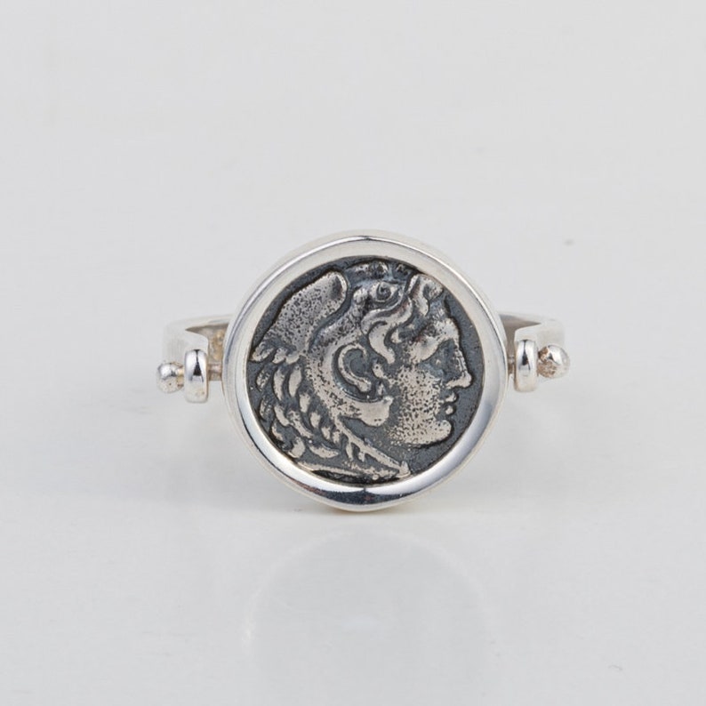 May include: A silver ring with a round coin-shaped design featuring a profile of a person's head with a laurel wreath. The ring is open and has a hinged design.