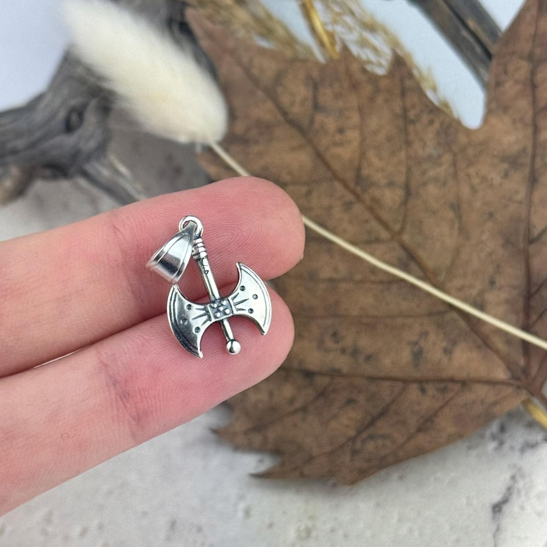 K&ouml;nnte beinhalten: Silberner Doppelbeil-Anh&auml;nger mit detailliertem Design. Die Axt hat oben eine Schlaufe zum Anbringen an einer Kette oder Schnur.