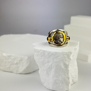 May include: A silver and gold ring with a vintage coin design and two red gemstones. The ring is displayed on a white rock-like surface.