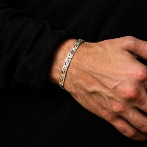 May include: A silver and gold bracelet with a geometric pattern. The bracelet is on a person's wrist.