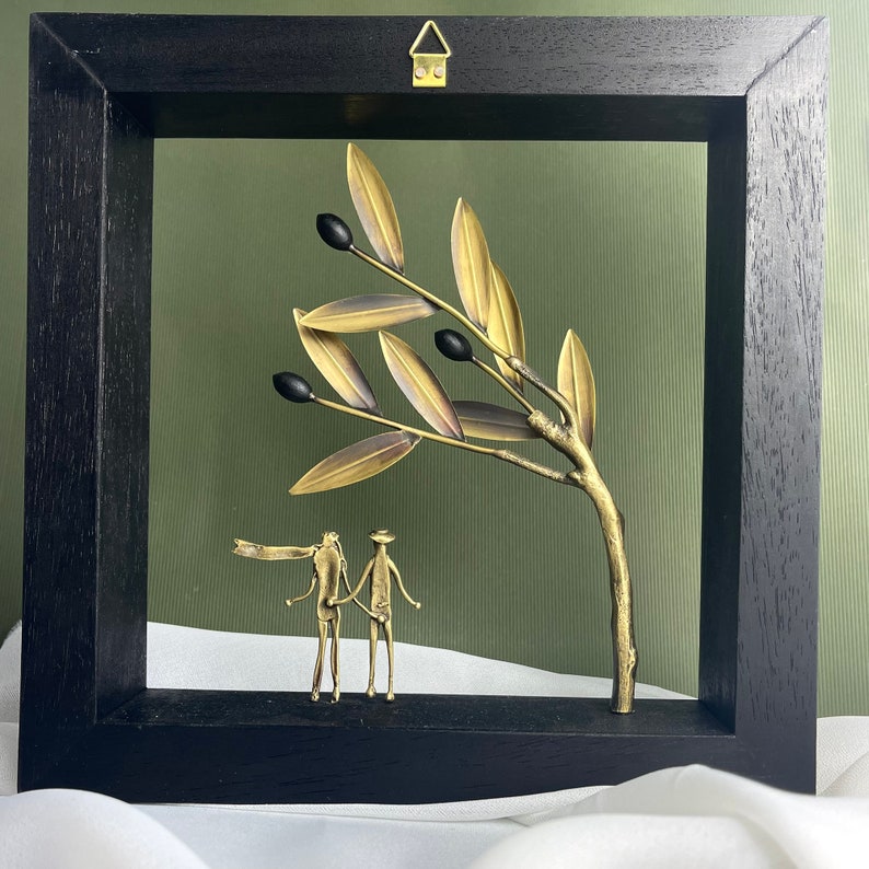 May include: A black framed wall art piece featuring a gold metal olive branch with black olives and two gold metal figures standing beneath it.