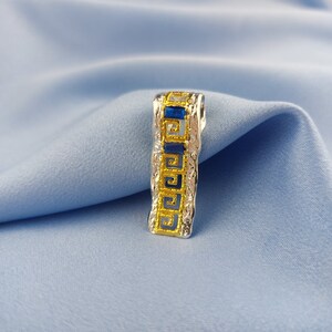 Peut inclure: Un pendentif en argent avec un motif de cl&eacute; grecque dor&eacute;e et des pierres pr&eacute;cieuses bleues.