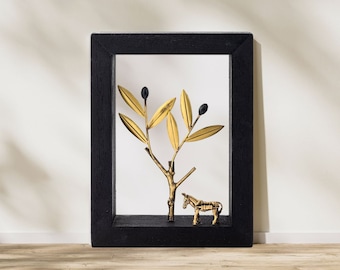 Op de afbeelding: Een zwarte geframed schaduwdoos met een gouden metalen olijftak en een gouden metalen ezelbeeldje erin.