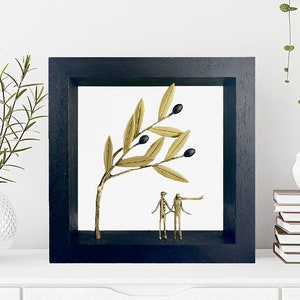 May include: A black framed shadow box with a gold olive branch and black olives. Two gold figures stand beneath the branch.