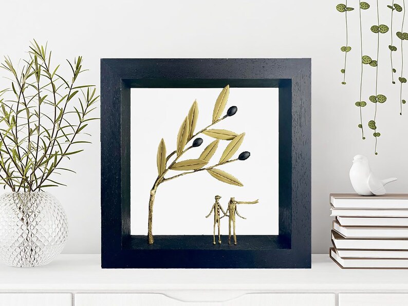 May include: A black framed shadow box with a gold olive branch and black olives. Two gold figures stand beneath the branch.