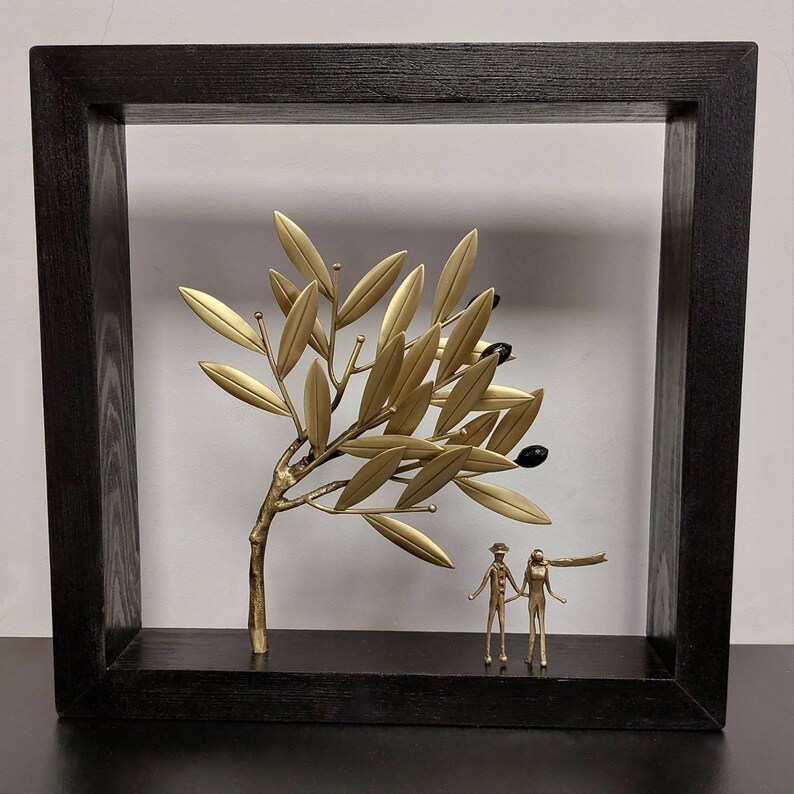 May include: A gold metal sculpture of an olive tree with black olives, framed in a black wood frame. Two gold metal figures stand beneath the tree.