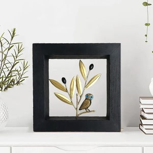 May include: A black framed wall decor with a gold metal olive branch and a small owl with blue eyes. The owl is perched on the branch.