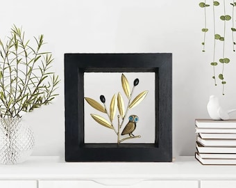 May include: A black framed wall decor with a gold metal olive branch and a small owl with blue eyes. The owl is perched on the branch.
