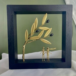 May include: A black framed wall art piece featuring a gold metal sculpture of an olive branch with black olives and two figures standing beneath it.